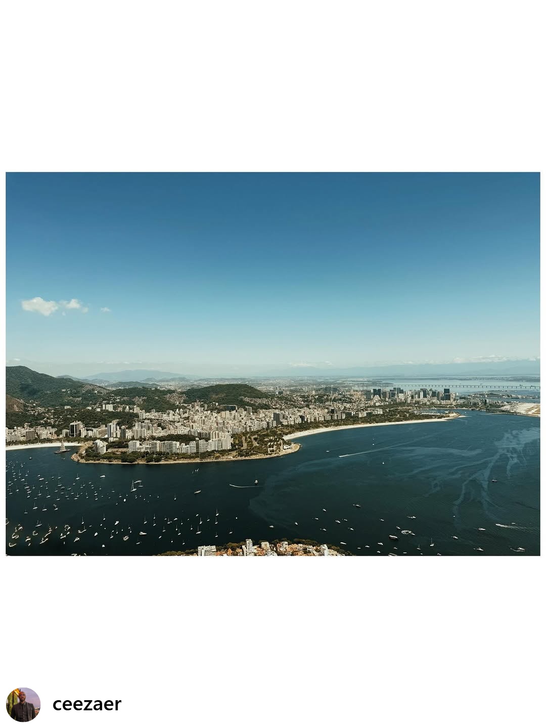 Event Coverage - Aerial view of Guanabara Bay with hundreds of sailboats, coastal cityscape, forested mountains, and a long bridge stretching across the water under clear blue skies