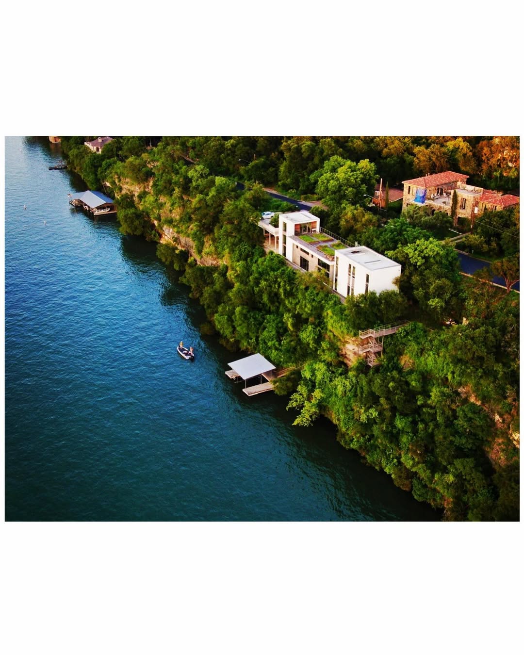 Real Estate Aerial - Aerial drone view of luxury waterfront properties along a lake with modern white architecture, boat docks, and lush green trees